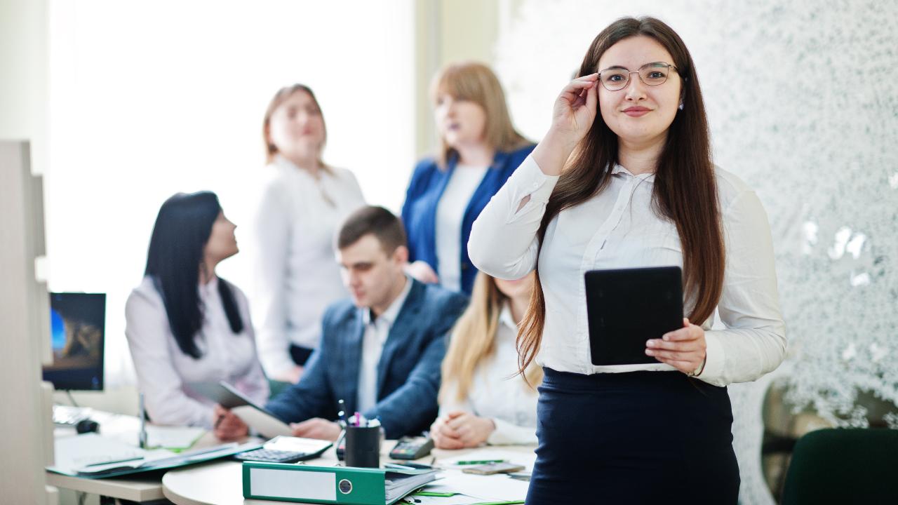 Giovane donna impiegata di banca con, sullo sfondo, alcuni colleghi.